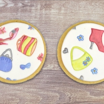 Dois biscoitos redondos decorados com desenhos coloridos de acessórios de moda, sobre uma superfície de madeira