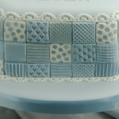 Barra decorativa retangular com padrão de quadrados azuis e creme com várias texturas, sobre superfície branca com bordado rendado.