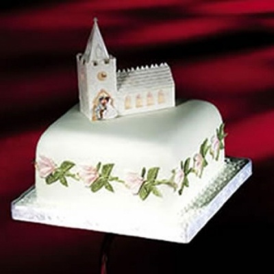 Bolo branco decorado com flores cor-de-rosa, folhas verdes e igreja branca no topo