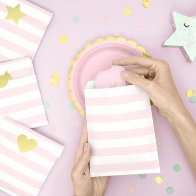 Sacos de papel rosa e branco com decorações douradas e mãos a colocar objeto rosa dentro, sobre fundo rosa com confetes e estrela decorativa.