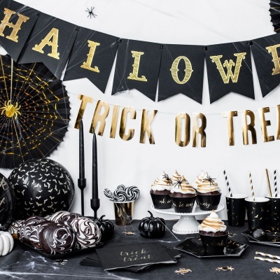 Decoração de Halloween com bandeiras, balões, cupcakes, velas e copos em preto e dourado sobre mesa preta