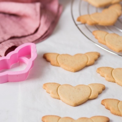 Biscoitos em forma de coração com asas, molde rosa e ovos numa tigela ao fundo