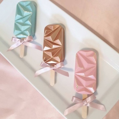 Três picolés decorativos azul, castanho e rosa com padrão geométrico e laços rosa em travessa branca.