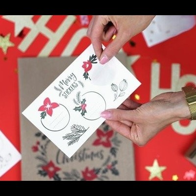 Folha de adesivos natalícios sendo segurada por mãos com relógio dourado, fundo decorado com motivos de Natal.