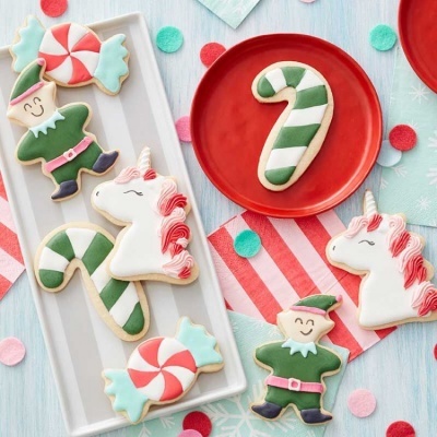 Bolachas decoradas de Natal em forma de duende, unicórnio, bengala e caramelos sobre pratos e superfície azul clara.
