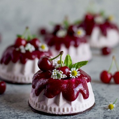 Pequeno bolo decorado com cerejas, flores e folhas verdes sobre superfície cinzenta