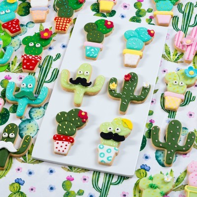 Biscoitos decorados em forma de cactos coloridos sobre tabuleiros e pano com estampa de cactos.