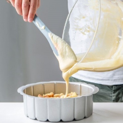 Forma de metal para bolo com massa amarela sendo despejada numa superfície branca.