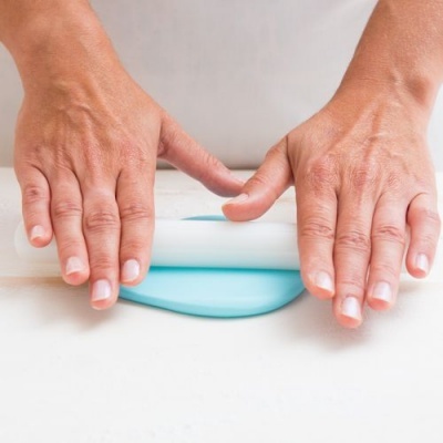 Duas mãos a pressionar massa azul clara com um rolo branco sobre superfície branca