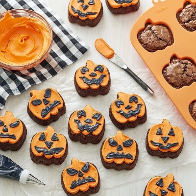 Muffins decorados de Halloween com cobertura alaranjada e decoração preta em forma de abóboras, utensílios de cozinha ao lado.
