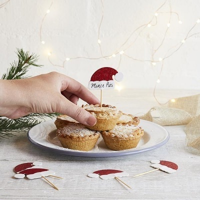 Quatro tartes pequenas num prato com decoração de Natal e mão segurando etiqueta 'Mince Pies'.