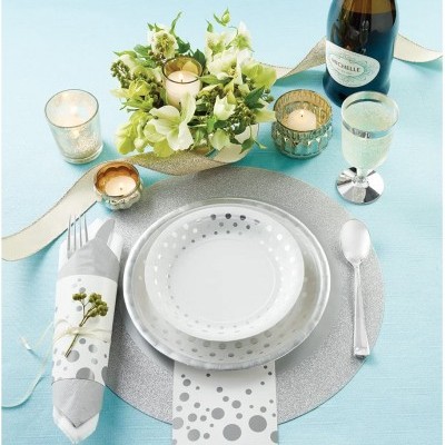 Mesa posta com pratos brancos de padrão de círculos cinzentos, guardanapo com padrão a combinar, velas, arranjo floral verde, copo de água, garrafa e talheres sobre toalha azul claro.