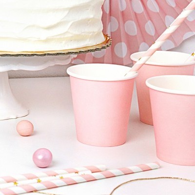 Copos de festa cor-de-rosa com canudos listrados e bolo branco ao fundo