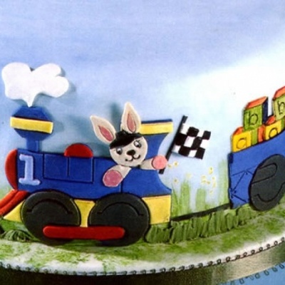 Bolo decorado com comboio azul e coelho com bandeira quadriculada transportando blocos coloridos.