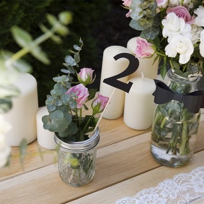Arranjos florais em frascos de vidro com flores brancas e rosas e rótulo número 2 sobre mesa de madeira com renda e velas brancas