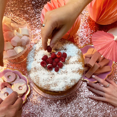 Bolo redondo com açúcar em pó e framboesas decorado, rodeado por vários doces cor-de-rosa numa mesa brilhante cor-de-rosa