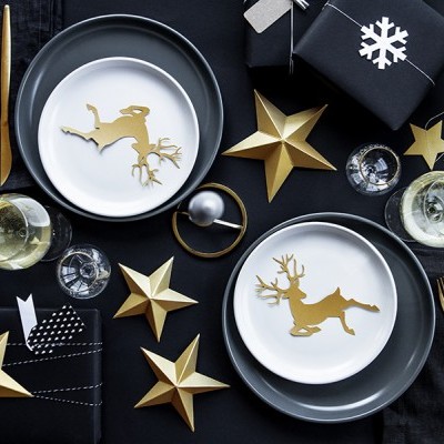 Mesa de jantar decorada para Natal com pratos e talheres em dourado e cinzento, estrelas decorativas e caixas de presente negras.