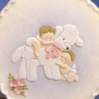 Bordado de ursinho branco com laço rosa e dois bebés a abraçá-lo em tecido redondo com flores cor-de-rosa.