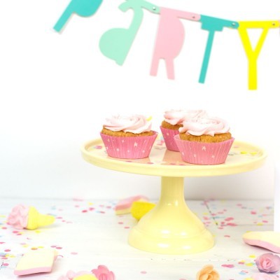Três cupcakes cor-de-rosa sobre suporte amarelo claro numa mesa branca com decoração de festa colorida