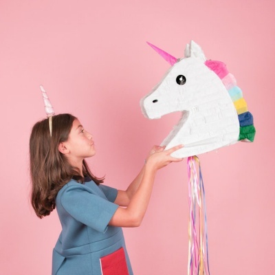 Menina com túnica azul segura piñata de unicórnio colorido em fundo rosa