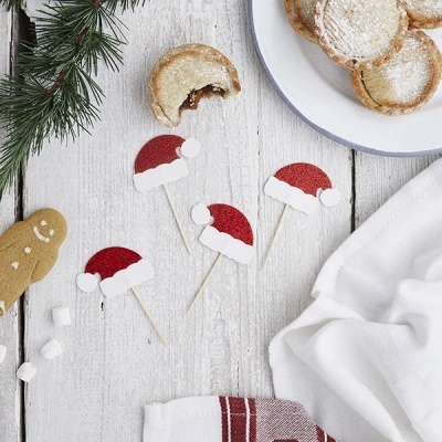 Pequenas decorações de chapéu de Pai Natal em palitos sobre mesa com biscoitos e pano branco