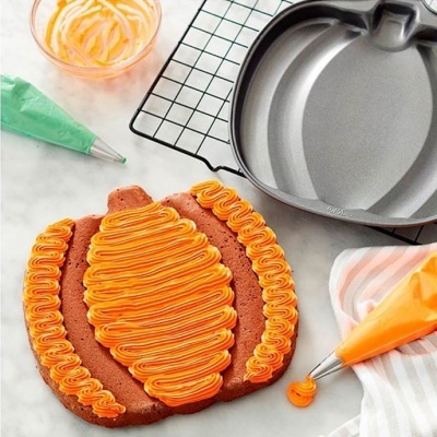 Forma metálica para bolo em formato de abóbora com bolo decorado e utensílios de confeitaria à volta