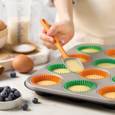 Tabuleiro metálico para muffins com formas coloridas e massa sendo colocada com colher