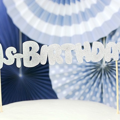 Topo de bolo '1st BIRTHDAY' em glitter prateado sobre bolo branco com decoração azul e branca ao fundo