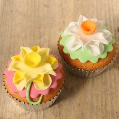 Dois cupcakes com decoração floral em glacê, um com fundo rosa e flor amarela, outro com fundo verde e flor branca e laranja.