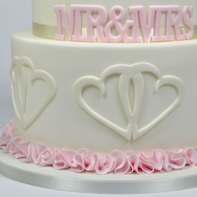 Bolo de casamento branco com corações entrelaçados em relevo e volantes rosa na base, com topo rosa MR & MRS