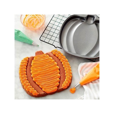 Forma metálica cinzenta de abóbora com bolo decorado com glacê laranja e ferramentas de decoração coloridas