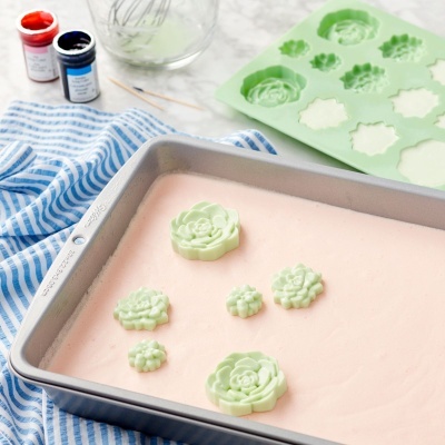 Forma de silicone verde com moldes de flores, tigela metálica com massa rosa e utensílios de cozinha numa bancada.