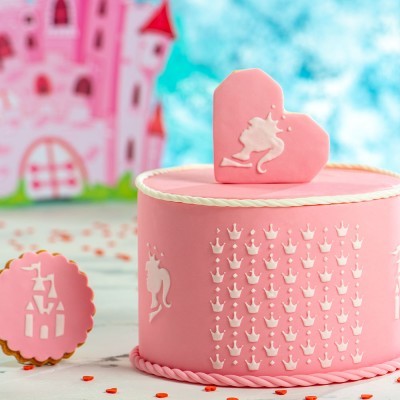 Bolo rosa com decoração de coroas e unicórnios, biscoitos decorados e castelo rosa ao fundo