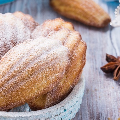 Três madeleines com açúcar em pó numa tigela azul clara sobre mesa de madeira com especiarias ao redor