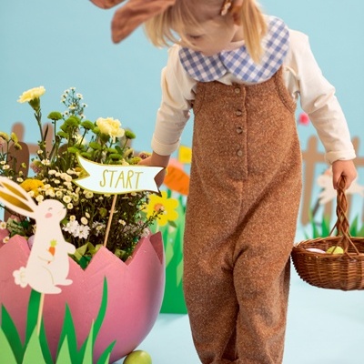 Criança com macacão castanho e camisa com gola quadriculada segurando cesta em cenário decorado