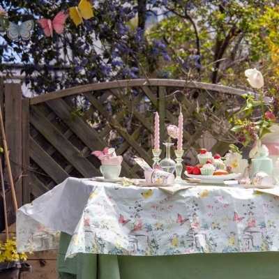 Mesa exterior com toalha decorada, velas cor-de-rosa, chávenas, cupcakes e flores ao fundo