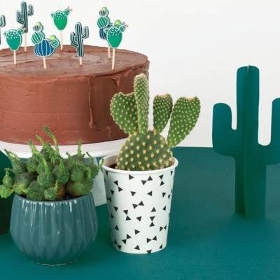 Bolo de chocolate com toppers de cactos, dois vasos com cactos reais e figura decorativa de cacto numa mesa verde.