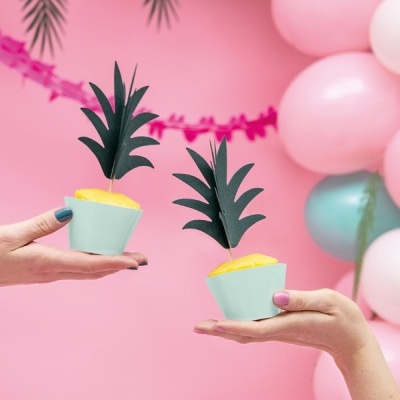 Duas mãos seguram cupcakes decorados com folhas verdes artificiais em fundo rosa com balões