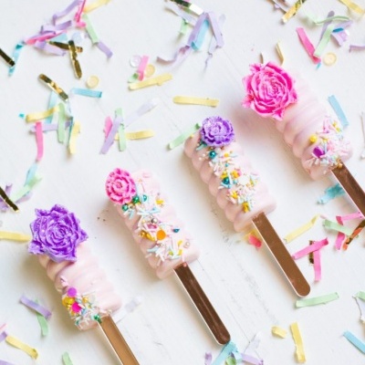 Três gelados cor-de-rosa claros decorados com flores de açúcar e confeitos coloridos rodeados por confetes multicoloridos