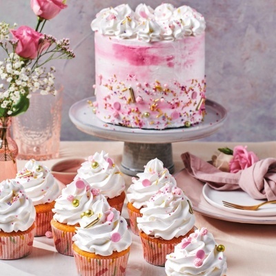 Bolo rosa e branco decorado com cupcakes semelhantes em mesa com flores e guardanapos rosa