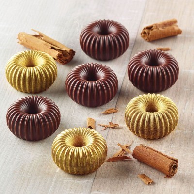 Bombons em forma de donut com ranhuras em chocolate castanho e dourado, com paus de canela ao redor, sobre superfície de madeira clara.
