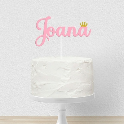 Bolo branco com topo de bolo cor-de-rosa escrito Joana e coroa amarela