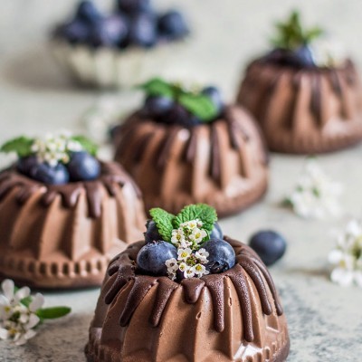Quatro mini bolos de chocolate decorados com mirtilos, hortelã e flores em fundo cinzento