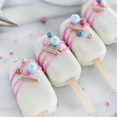 Gelados em palito com chocolate branco e decorações coloridas, sobre superfície branca com granulados.