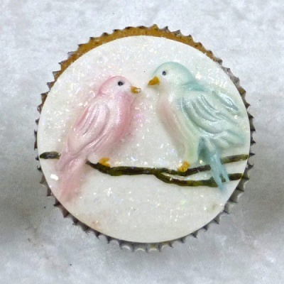 Cupcake com decoração de dois pássaros em glacê rosa e azul sobre glacê branco.