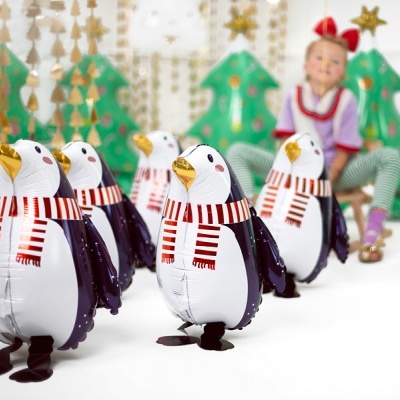 Cinco pinguins de peluche com cachecóis vermelhos e brancos alinhados; criança e decoração de Natal ao fundo