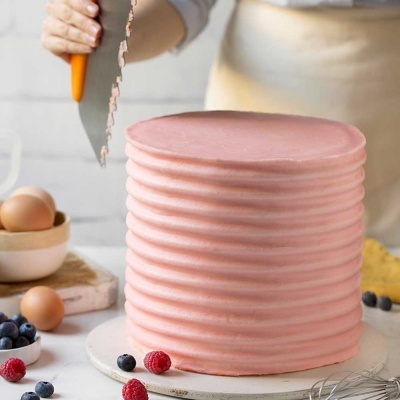 Bolo rosa alto com textura ondulada e faca preparada para cortar, frutas e ovos ao redor