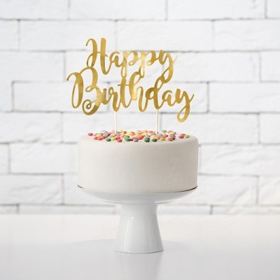 Bolo branco com confeitos coloridos e topo dourado 'Happy Birthday' em suporte branco