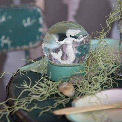 Globo de neve com sereia branca e base verde sobre mesa escura com plantas secas e concha