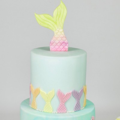 Bolo de dois andares azul claro com decoração de caudas de sereia coloridas.